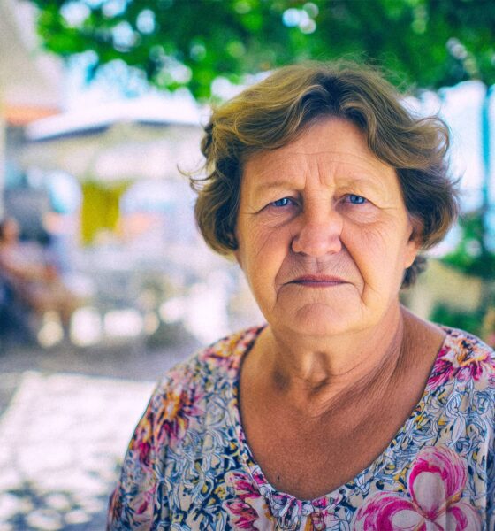 **SEO alt opis:** Starija žena ozbiljnog izraza lica stoji na terasi okruženoj cvijećem, s pogledom prema moru i ljetnoj mediteranskoj pozadini.
