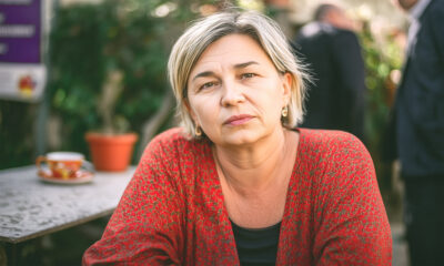 Zabrinuta žena srednjih godina u crvenom kardiganu sjedi za stolom na terasi kafića, s ozbiljnim izrazom lica i šalicom kave u pozadini.