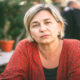 Zabrinuta žena srednjih godina u crvenom kardiganu sjedi za stolom na terasi kafića, s ozbiljnim izrazom lica i šalicom kave u pozadini.