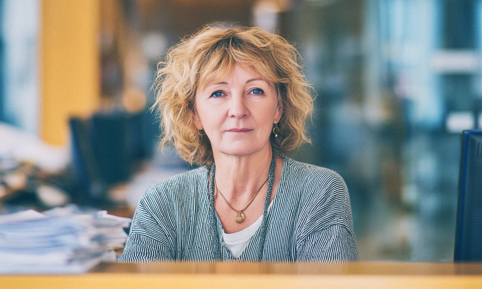 Sredovječna žena ozbiljnog izraza lica sjedi za uredskim stolom okružena papirima i računalima.