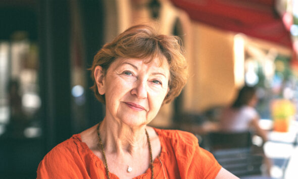 Žena srednjih godina s blagim osmijehom, u svijetloj majici s uzorkom, stoji na otvorenom; u pozadini hrvatska zastava i zgrade.