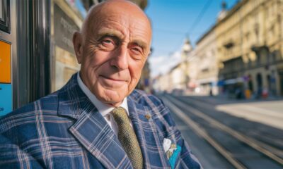 Stariji gospodin u kariranom odijelu i kravati stoji na ulici s tramvajskim tračnicama u pozadini, sunčan dan u gradu.