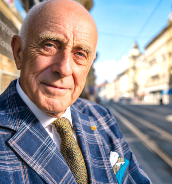 Elegantan stariji muškarac u plavom kariranom odijelu stoji uz tramvajsku stanicu na sunčanoj gradskoj ulici, s blagim osmijehom i prirodnim izrazom lica.