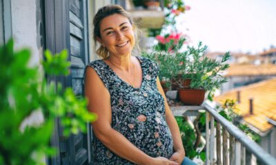 Trudnica nasmiješenog lica stoji na balkonu s cvjetnim lončanicama i pogledom na grad.