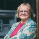 Starija žena s naočalama i crvenim ružem stoji ispred bankomata, odjevena u cvjetni sako.