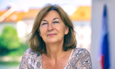 Žena srednjih godina s blagim osmijehom, u svijetloj majici s uzorkom, stoji na otvorenom; u pozadini hrvatska zastava i zgrade.