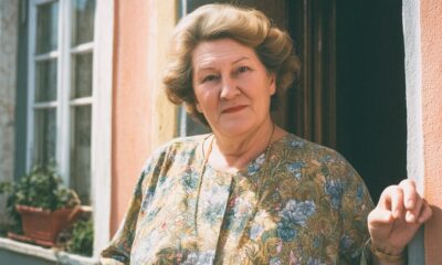 Starija žena sa staromodnom frizurom stoji na vratima obiteljske kuće.