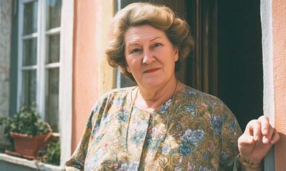Starija žena sa staromodnom frizurom stoji na vratima obiteljske kuće.
