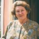 Starija žena sa staromodnom frizurom stoji na vratima obiteljske kuće.