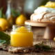 Staklenka lemon curda ili kreme od limuna na drvenom srustikalnom stolu, iza nje u pozadini Pavlova i nekoliko svježih limuna.