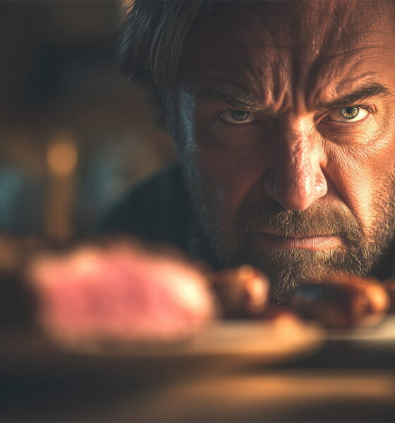 Bijesan muškarac za stolom s kobasicama i T-bone odreskom, simbol unutarnjeg sukoba mesojeda prema vegetarijanstvu.