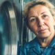 Žena u kasnim pedesetima stoji uz veliki industrijski stroj za pranje rublja u tvornici, pod fluorescentnim svjetlom. Na licu joj se vidi umor, ruke su grube od rada, a izraz dostojanstven. Dokumentaristička fotografija dobro osvijetljenog radnog prostora.
