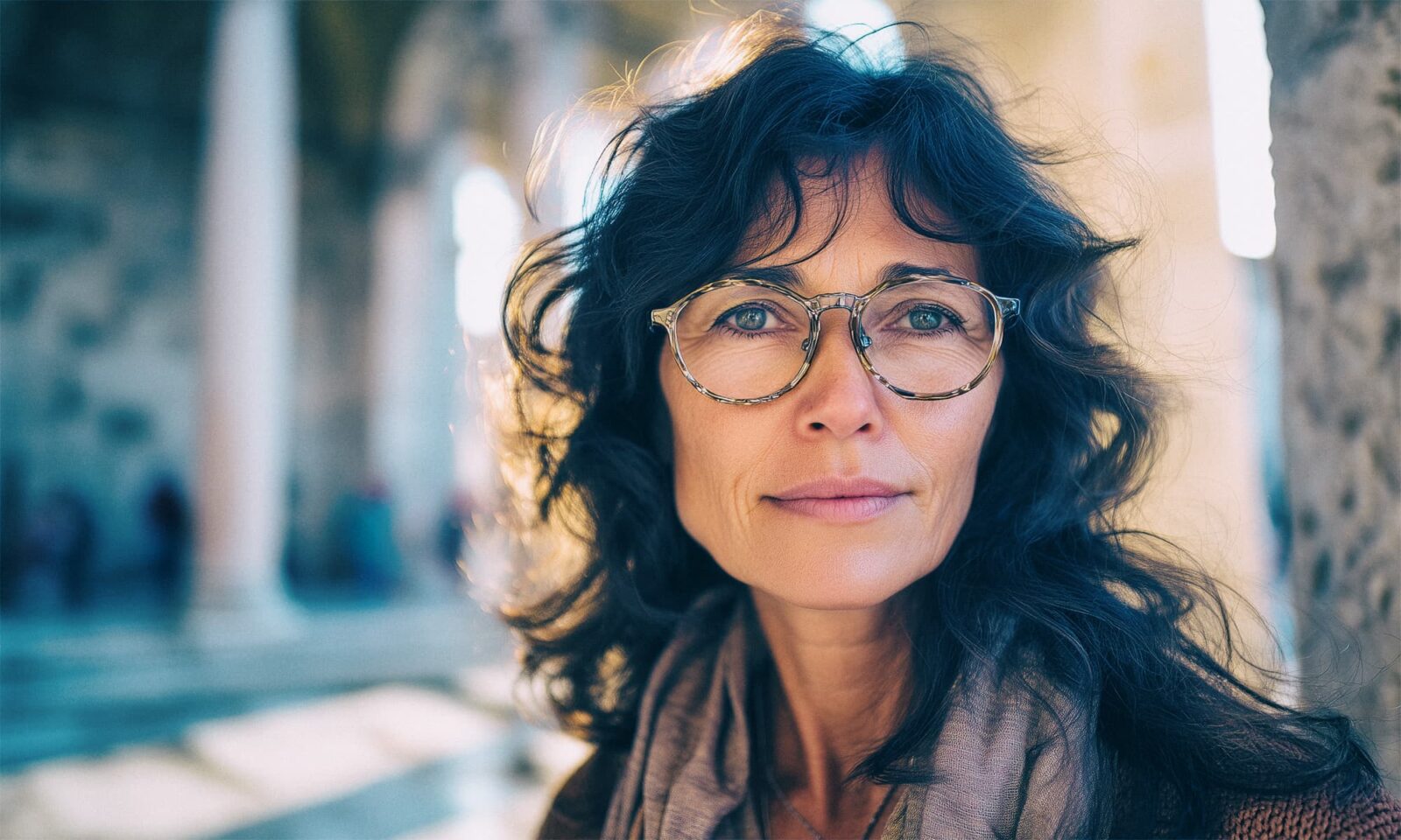 Portret žene srednjih godina s naočalama, stoji na Peristilu u Splitu u sunčanom jesenjem danu, gleda izravno u kameru s blagim osmijehom, u pozadini su kameni stupovi i povijesna arhitektura.