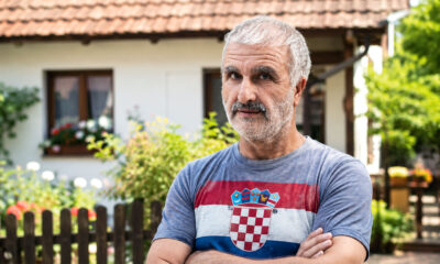 Muškarac u majici s hrvatskom zastavom stoji u dvorištu obiteljske kuće i gleda prema kameri. Svijetla, prirodna fotografija s urednim bojama i atmosferom domaćeg okruženja.