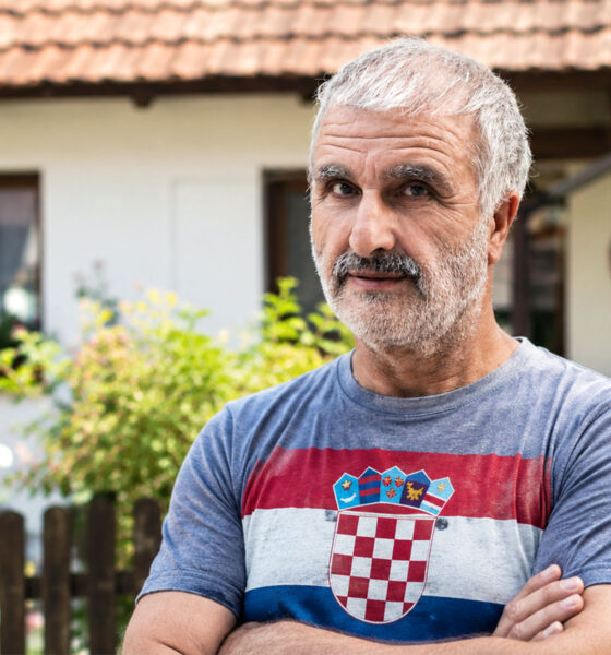 Muškarac u majici s hrvatskom zastavom stoji u dvorištu obiteljske kuće i gleda prema kameri. Svijetla, prirodna fotografija s urednim bojama i atmosferom domaćeg okruženja.