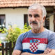 Muškarac u majici s hrvatskom zastavom stoji u dvorištu obiteljske kuće i gleda prema kameri. Svijetla, prirodna fotografija s urednim bojama i atmosferom domaćeg okruženja.