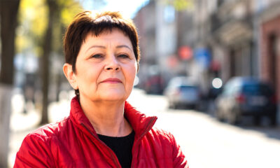 Žena u crvenoj jakni stoji na gradskoj ulici i samouvjereno gleda u kameru, s blagim zadovoljnim izrazom lica i prirodnim dnevnim svjetlom u kadru.