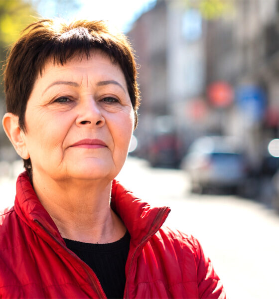 Žena u crvenoj jakni stoji na gradskoj ulici i samouvjereno gleda u kameru, s blagim zadovoljnim izrazom lica i prirodnim dnevnim svjetlom u kadru.