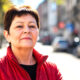 Žena u crvenoj jakni stoji na gradskoj ulici i samouvjereno gleda u kameru, s blagim zadovoljnim izrazom lica i prirodnim dnevnim svjetlom u kadru.