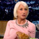 Helen Mirren u ružičastoj majici tijekom gostovanja u televizijskoj emisiji, s mikrofonom i zdjelom čipsa ispred sebe.