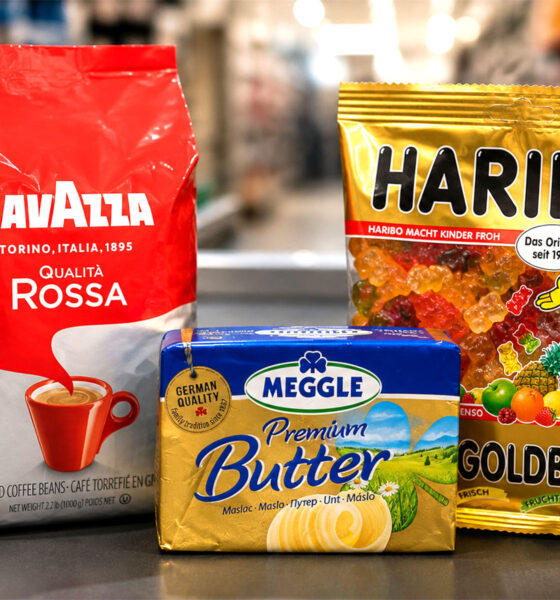 Na blagajni supermarketa uz zamućenu pozadinu trgovine stoje tri proizvoda čije se cijene uspoređuju: Lavazza Qualità Rossa kava u zrnu, Haribo Zlatni medo gumeni bomboni i Meggle Premium maslac.