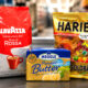 Na blagajni supermarketa uz zamućenu pozadinu trgovine stoje tri proizvoda čije se cijene uspoređuju: Lavazza Qualità Rossa kava u zrnu, Haribo Zlatni medo gumeni bomboni i Meggle Premium maslac.
