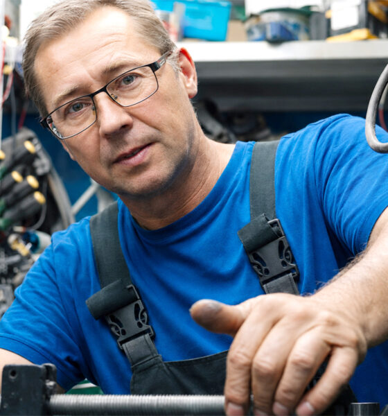 Automehaničar u plavoj radnoj odjeći popravlja vozilo u radionici i gleda u kameru.