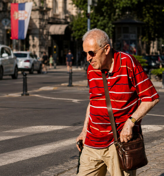 Stariji muškarac u crvenoj majici i sa smeđom torbom hoda gradskom ulicom tijekom sunčanog dana u Beogradu.