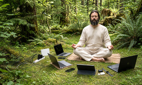 Bradati guru meditira u šumi okružen laptopima, mobitelima, routerom, punjačima i kabelima.