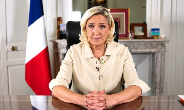 Marine Le Pen sjedi za radnim stolom u uredu, uz francusku zastavu u pozadini.