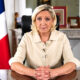 Marine Le Pen sjedi za radnim stolom u uredu, uz francusku zastavu u pozadini.