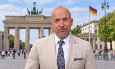 Alexander Hoffmann u svijetlom odijelu, s Brandenburškim vratima i njemačkom zastavom u fotomontiranoj pozadini.