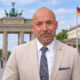 Alexander Hoffmann u svijetlom odijelu, s Brandenburškim vratima i njemačkom zastavom u fotomontiranoj pozadini.