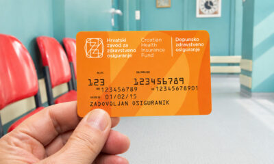 Ruka drži narančastu karticu HZZO-ova dopunskog zdravstvenog osiguranja u svijetloj bolničkoj čekaonici s crvenim stolicama.