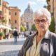 Starija žena sijede kose i naočala stoji na sunčanoj ulici talijanskoga grada, s povijesnim zgradama i prolaznicima u pozadini.