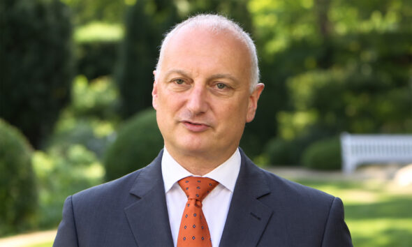 Njemački industrijalac Nikolas Stihl u tamnom odijelu i narančastoj kravati stoji na otvorenom, u sredini kadra, dok je iza njega zamućena zelena vrtna pozadina s prirodnim svjetlom.
