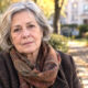 Starija žena sijede kose u tamnom kaputu i šarenom šalu, na klupi u jesenskom parku.