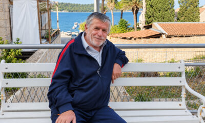Blagoje Babić sjedi na bijeloj klupi u Visu, s morem, palmama i kamenim kućama u pozadini.