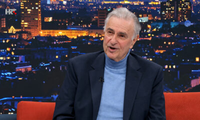 Goran Milić u televizijskom studiju, u tamnom sakou i plavoj dolčeviti, s panoramom grada u pozadini.