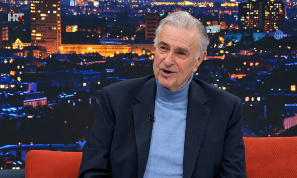 Goran Milić u televizijskom studiju, u tamnom sakou i plavoj dolčeviti, s panoramom grada u pozadini.