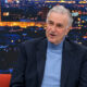Goran Milić u televizijskom studiju, u tamnom sakou i plavoj dolčeviti, s panoramom grada u pozadini.