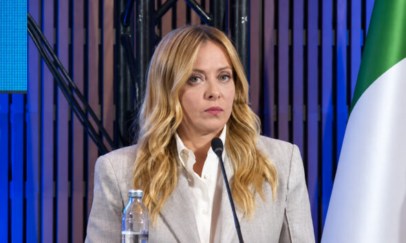 Giorgia Meloni u sivom sakou i bijeloj košulji pred mikrofonom, uz talijansku zastavu.