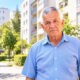 Stariji muškarac u plavoj košulji stoji ispred stambenih zgrada u sunčanom gradskom naselju.