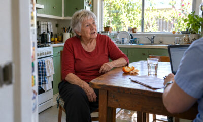 Starija žena u crvenoj majici sjedi za kuhinjskim stolom i razgovara s djelatnicom koja ispred sebe ima otvoren laptop i dokumente.